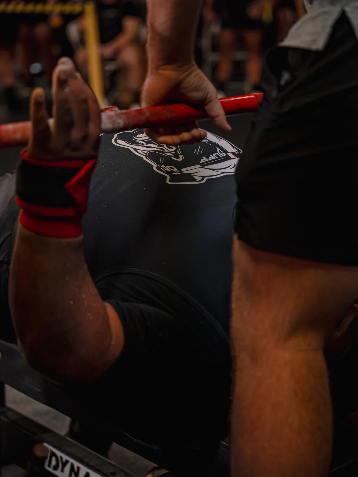Close-up of a lifter gripping a red barbell with a spotter assisting at the bench